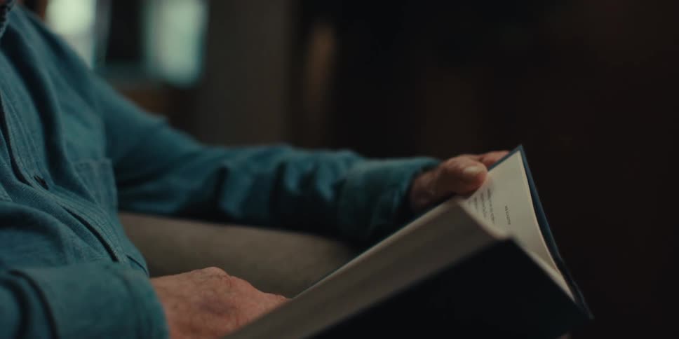 A happy elderly man wearing glasses and a blue shirt reads a book in the living room