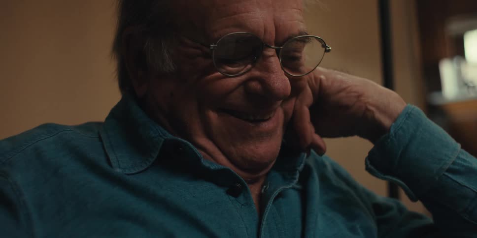 An elderly man in a blue shirt reads a book takes off his glasses and laughs in a dimly lit room