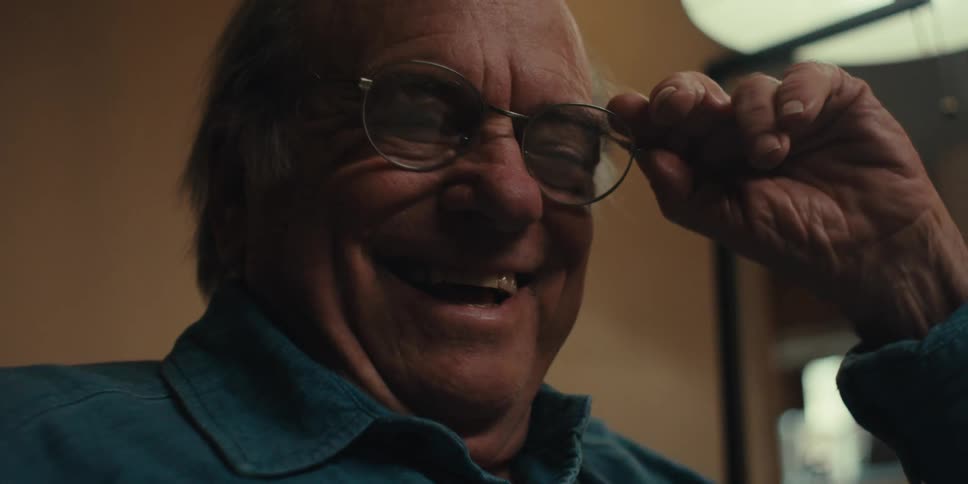 An elderly man wearing glasses and a blue shirt reads a book takes off his glasses and laughs in a dimly lit room