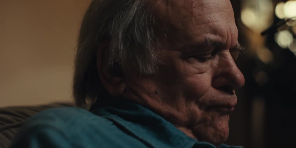 An elderly man with graying hair wearing a teal shirt thoughtfully putting on his glasses in a dimly lit room