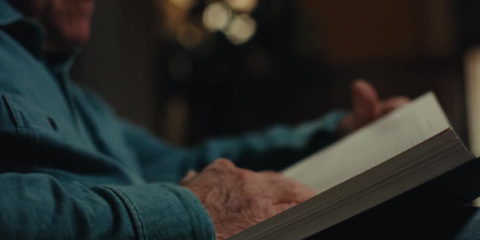 Elderly man wearing glasses and a blue shirt reads a book turning a page