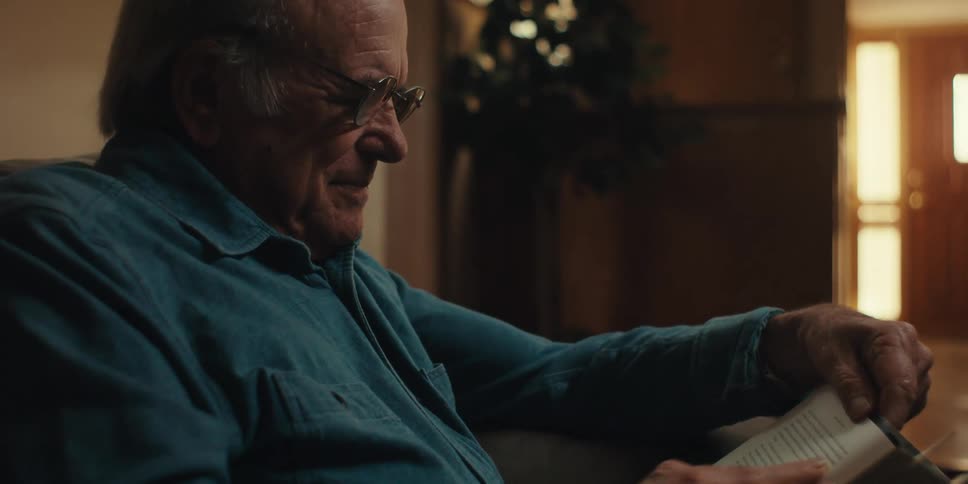 A happy elderly man wearing glasses and a blue shirt reads a book turning a page