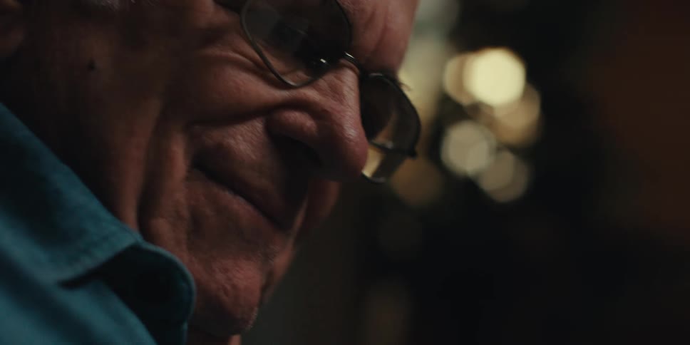 An elderly man with glasses and a blue shirt reads a book in a dimly lit room smiling
