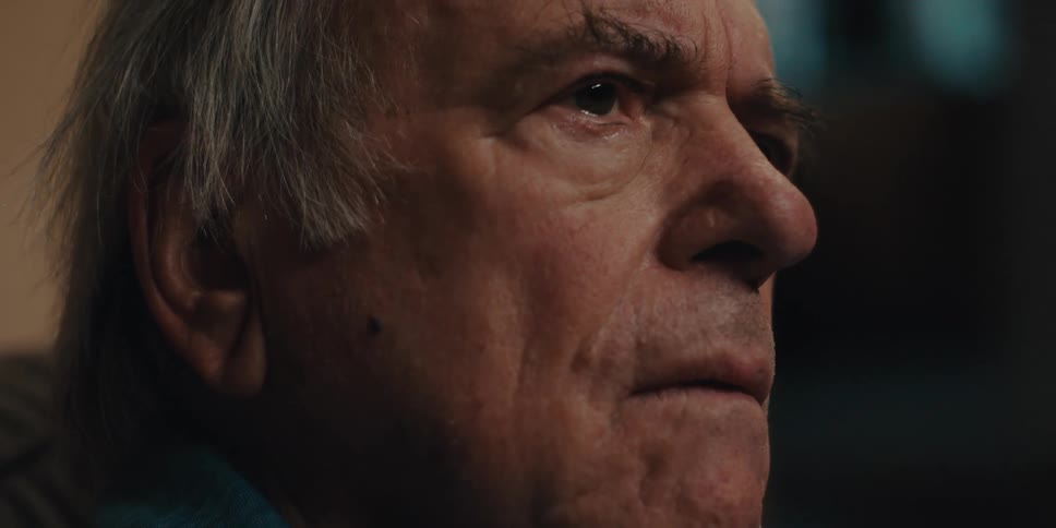 Close up of the face of an elderly man taking of his glasses on a dimly lit room