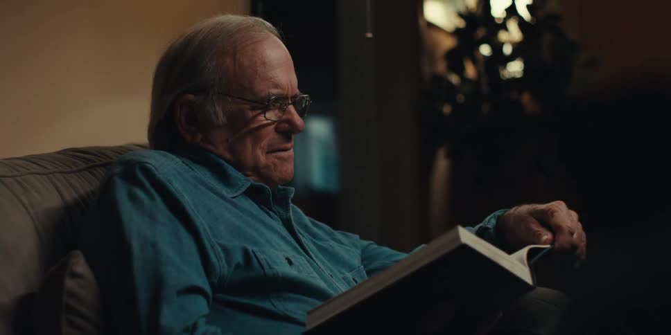 Elderly man wearing glasses and a blue shirt reads a book in a dimly lit room