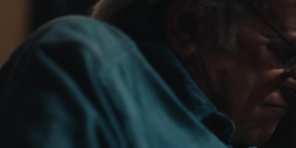 An elderly man wearing glasses and a blue shirt reads a book in a dimly lit room