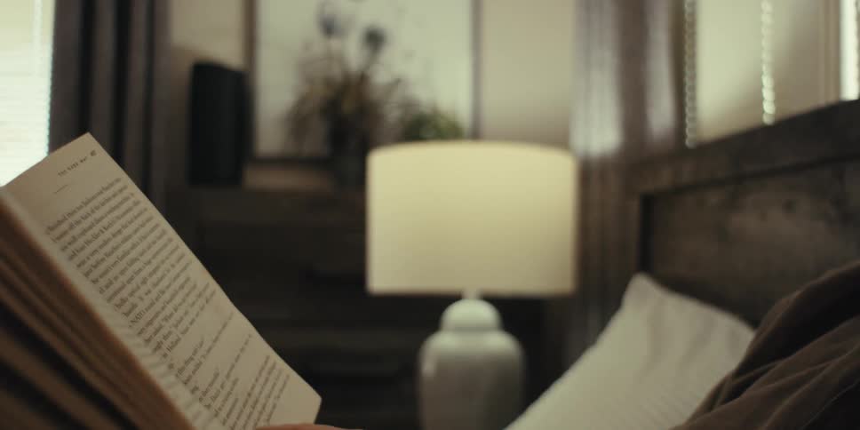 Elderly man wearing glasses and a brown shirt sits on the bed in the bedroom reading a book