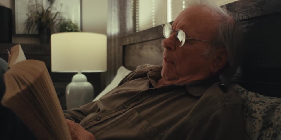 An older man reclining on a bed reading a book with focused attention he is wearing glasses and a brown shirt with a softly lit table lamp and potted plant in the background