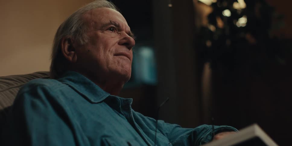 An elderly man in a blue shirt reading a book takes off his glasses and pauses thoughtfully
