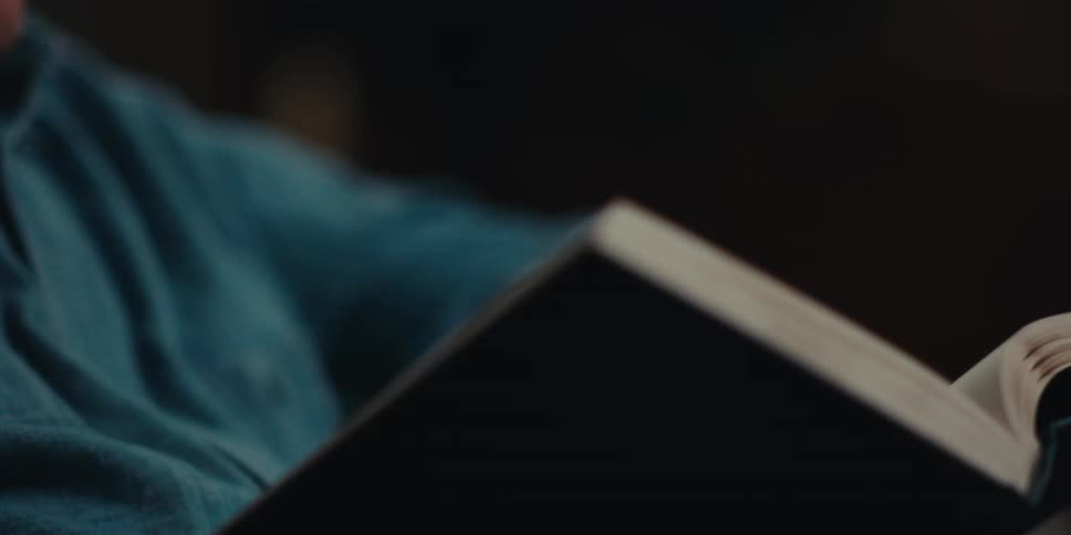 An elderly man wearing glasses and a blue shirt reads a book in a dimly lit room