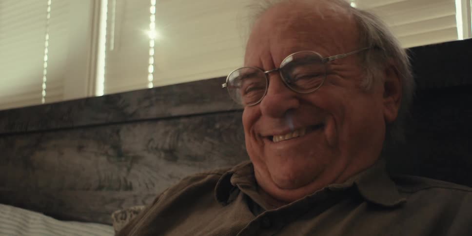 An elderly man sits on his couch reading something that makes him laugh