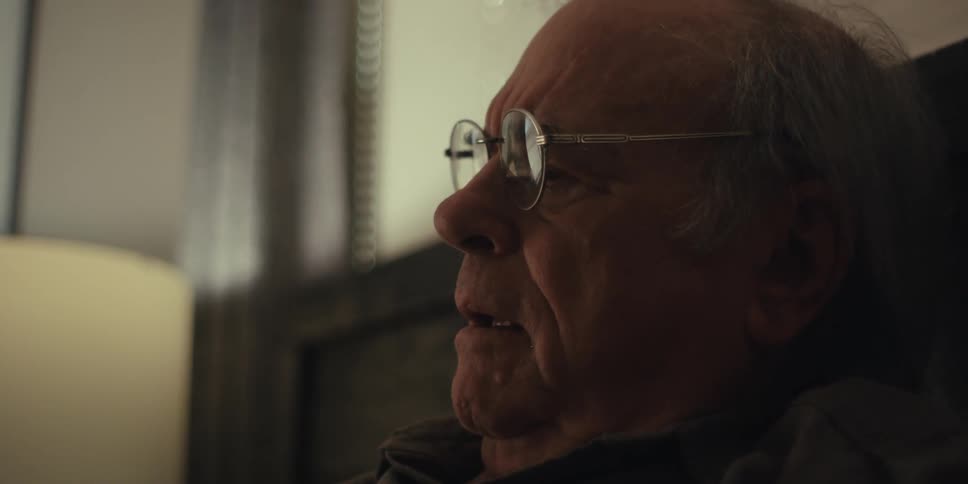 Sitting on the bed in the bedroom the elderly man with glasses reads a book and chuckles near the table lamp