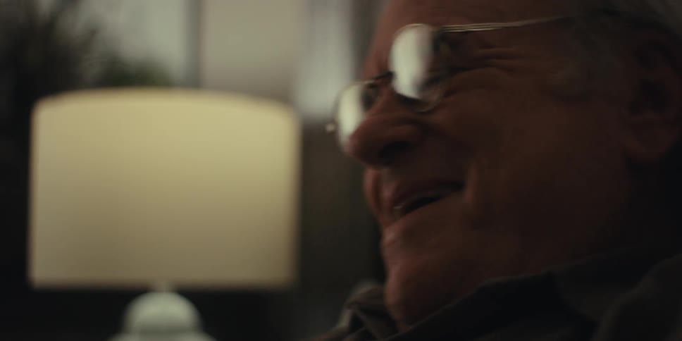 The elderly man wearing glasses laughs while sitting on the couch near a lamp