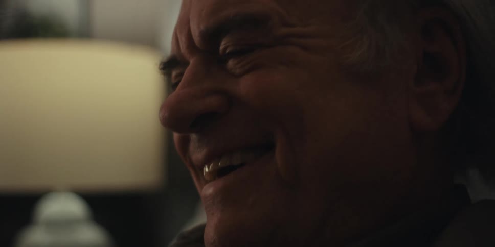 Elderly man takes off his glasses and laughs contently near a lamp in a dimly lit room