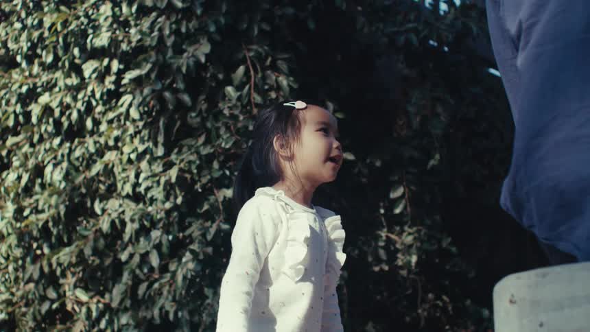 A little girl with a ponytail and a white shirt talks to her dad and then happily runs off to the playground