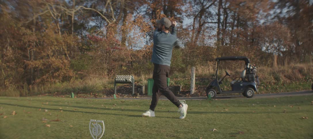 A man midswing while playing golf on a green field surrounded by autumncolored trees he is dressed casually in a blue sweater black pants and a cap with a golf cart and equipment visible in the background