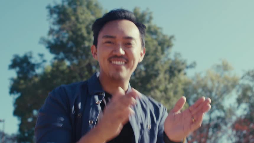 A man stands in a park smiling laughing and clapping his hands as the camera moves wildly up and down