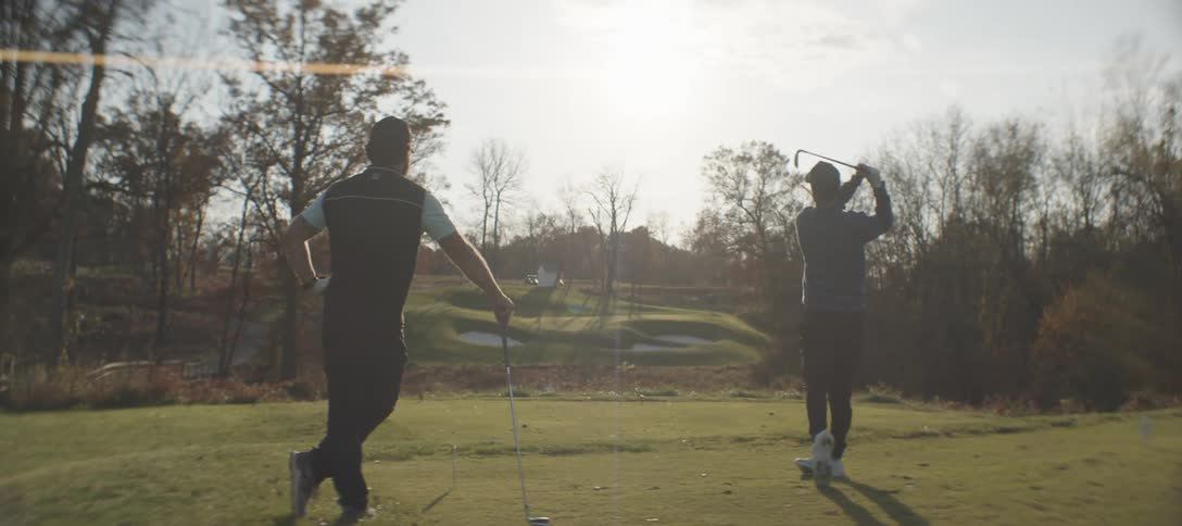 One golfer leans on his club while another takes a swing on a sunny autumn day