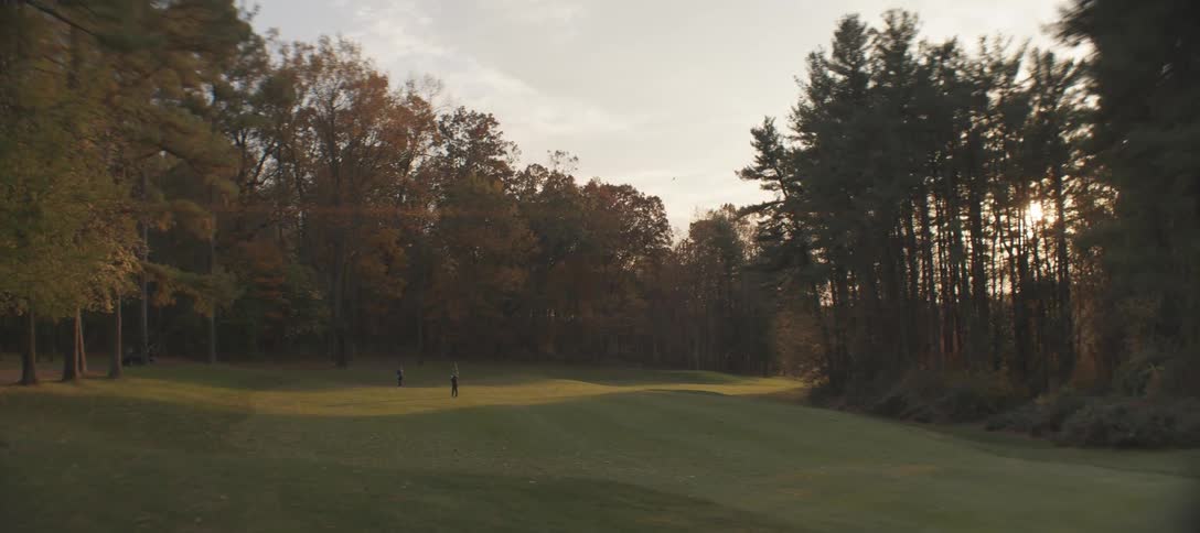 Distant two golfers playing on a golf course as the sun sets casting long shadows across the green grass