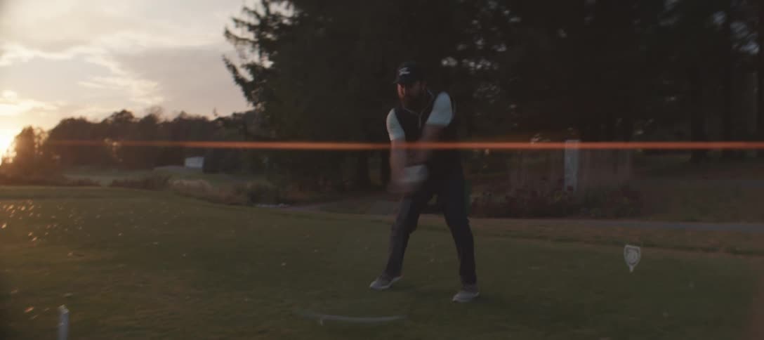 A golfer wearing a dark vest light shirt and cap strikes a golf ball with a golf club on the course at sunset