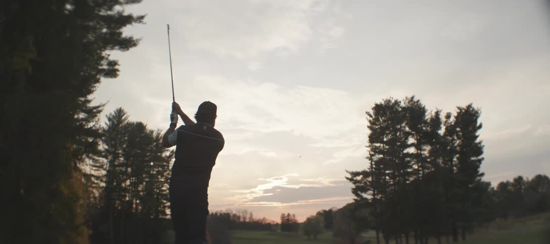 Golfer with a beard a cap a white shirt and black vest strikes golf ball with the club on the golf course at sunset