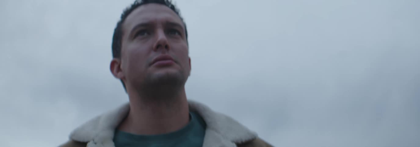 A man in a brown jacket with a white fleece collar stands impatiently under a cloudy sky
