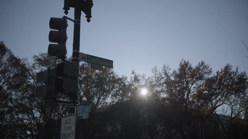 A traffic light and street signs at the intersection of constitution avenue nw in washington dc with the sun shining brightly behind bare trees