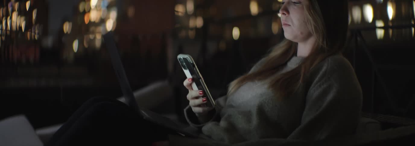 Woman with brown hair and a brown sweater uses smartphone and a laptop with illuminated tall buildings in the background