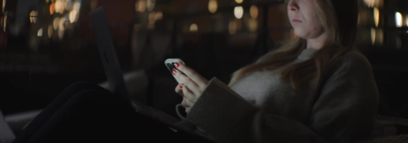 Woman with brown hair and a sweater uses a smartphone with a laptop on her lap with illuminated tall buildings in the background