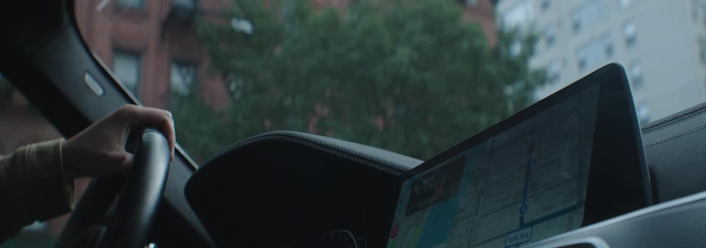 A person driving a car with a city street visible through the windshield and a large folded down dashboard screen displaying a map