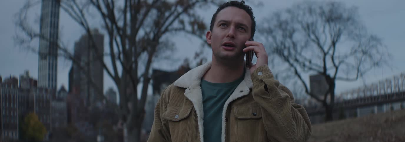 A man wearing a brown corduroy jacket talks on the phone with the city skyline in the background