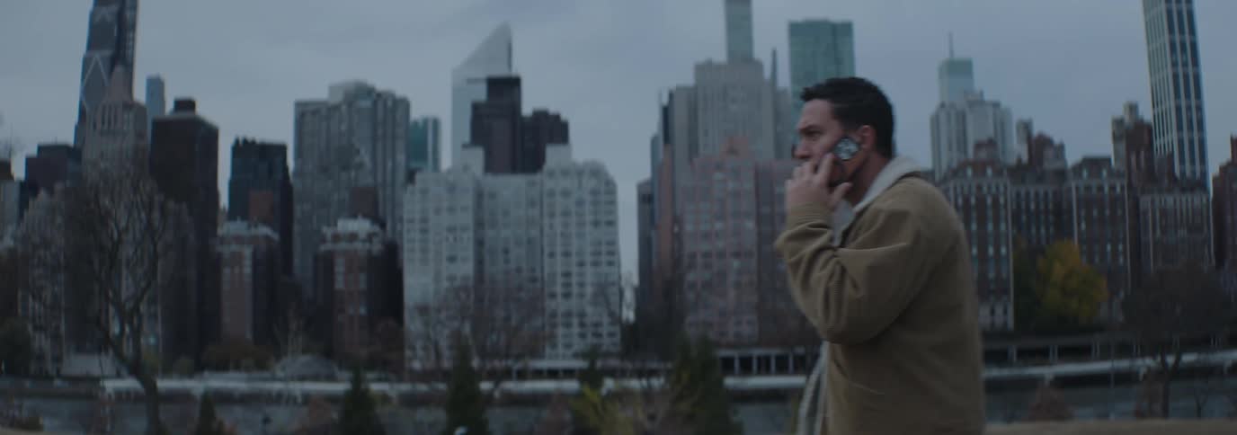 On a cloudy day a man wearing a brown corduroy jacket talks on the phone with the city skyline in the background