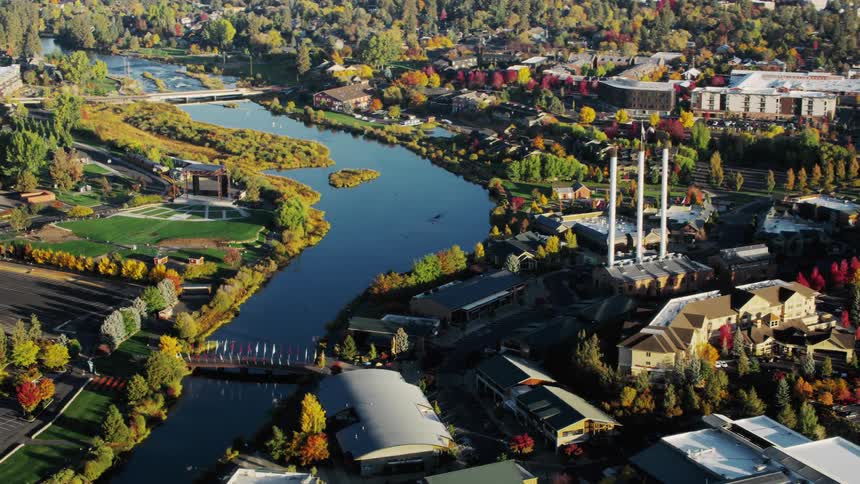 N aerial view of a city where a river winds through surrounded by lush greenery and buildings including a large industrial complex with smokestacks with autumn colors evident as many trees display vibrant shades of brown