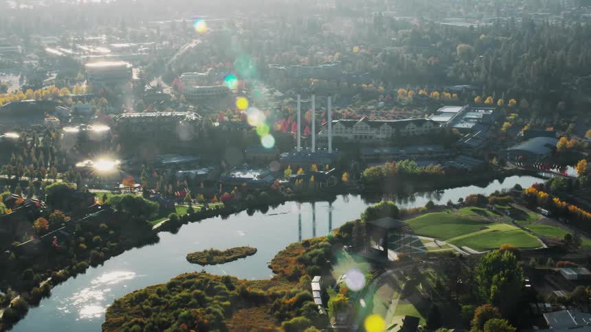 An aerial view of a city with a river winding through it surrounded by lush green trees and buildings including a large industrial complex with smokestacks