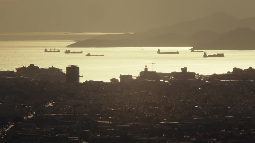 A coastal cityscape at sunset with several cargo ships anchored in the shimmering waters of a bay