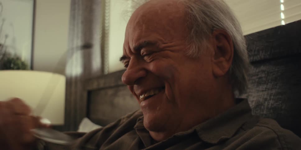 An elderly man wearing a brown shirt sits on a bed in the bedroom reading a book taking off his glasses and looking pensive