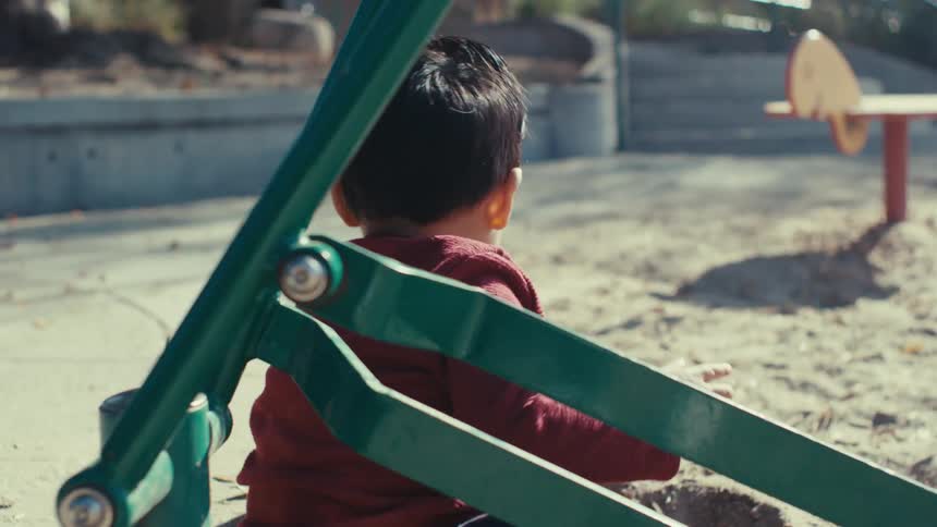 A toddler in a maroon shirt sits in a sandbox at a playground viewed from behind and partially obscured by playground equipment