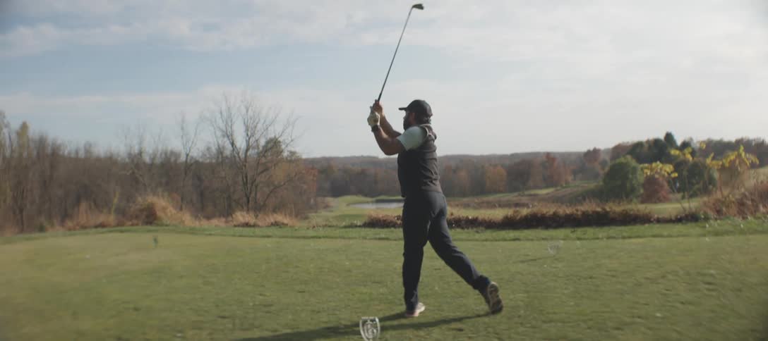 A golfer with a cap a beard a vest and a white tshirt striking the ball on a golf course