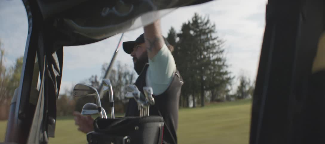 A man with a beard and a cap is standing outdoors next to a golf cart selecting golf clubs on the golf course