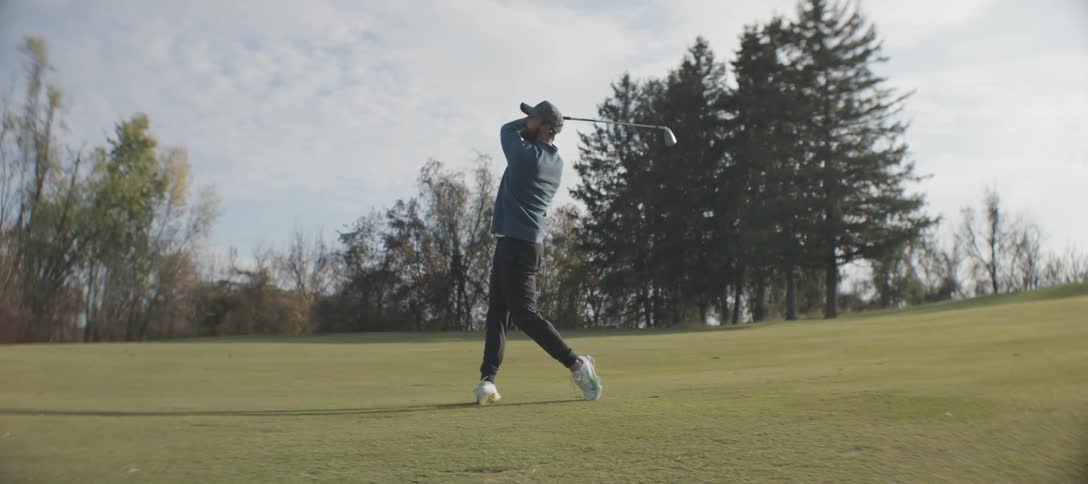 A golfer with a beard a cap a blue shirt and black pants strikes golf ball with a club on the course