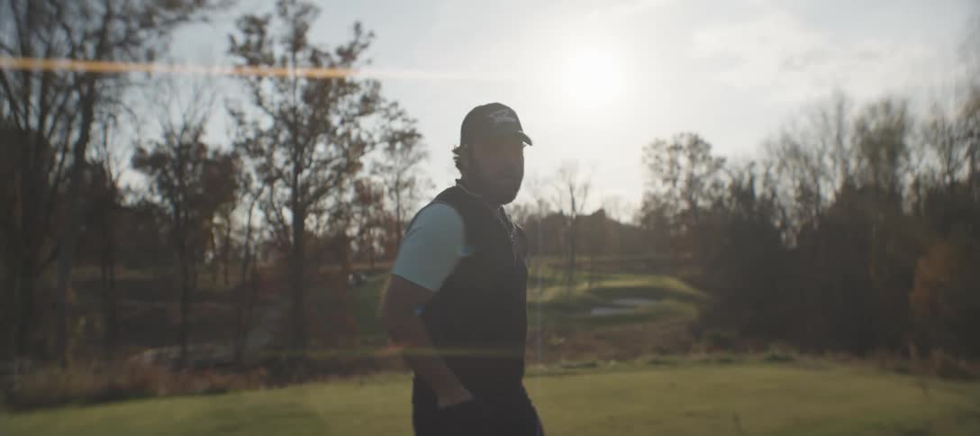 A golfer with a beard a black vest and a white shirt is silhouetted against the sun on the golf course