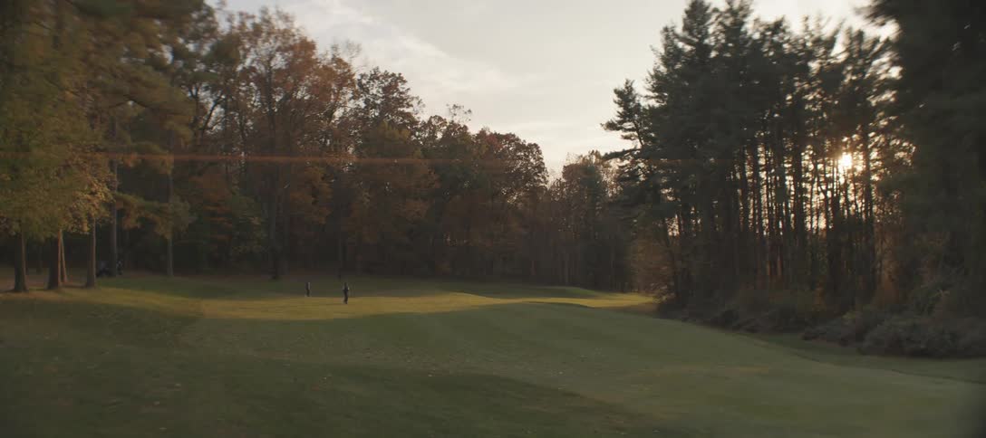 Two people stand on a golf course surrounded by autumnal trees under a partly cloudy sunset sky