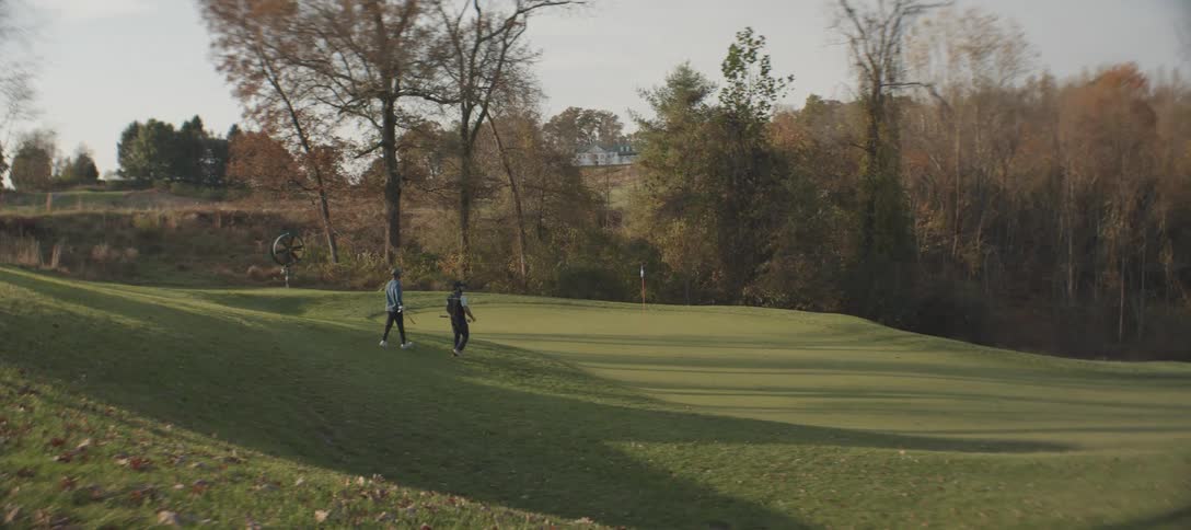 Two golfers walking along a fairway on a golf course with a wooded area and a hill in the background