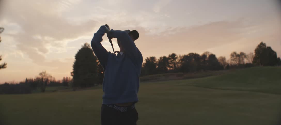 A golfer silhouetted against a sunset sky striking the ball on a golf course framed by distant trees