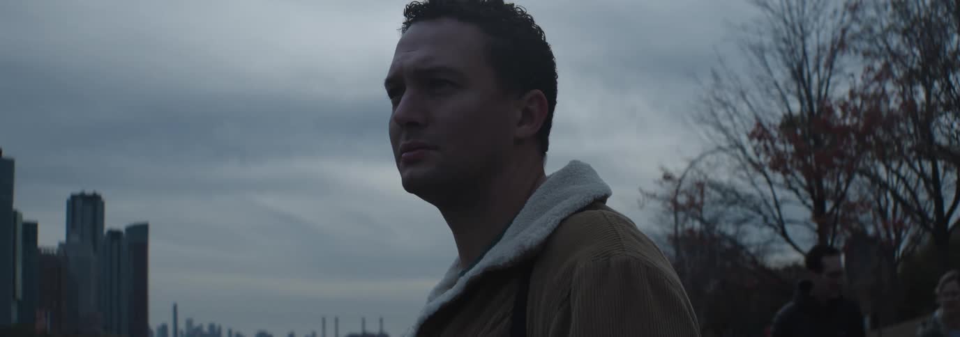 A man in a brown jacket with a white fleece collar stands impatiently under a cloudy sky in the city
