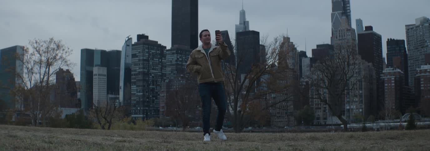A person stands in a park holding a phone with tall modern skyscrapers and leafless trees in the background