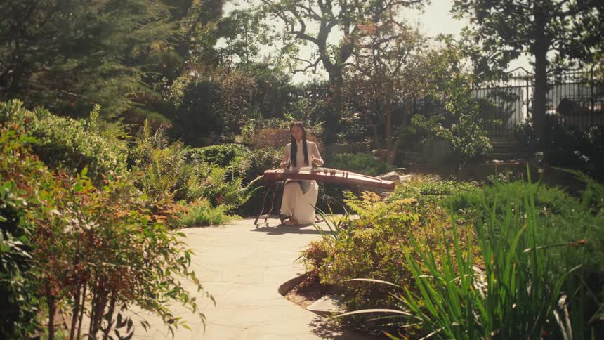 Woman plays a traditional stringed instrument sitting on a paved path in the middle of a lush sunny garden