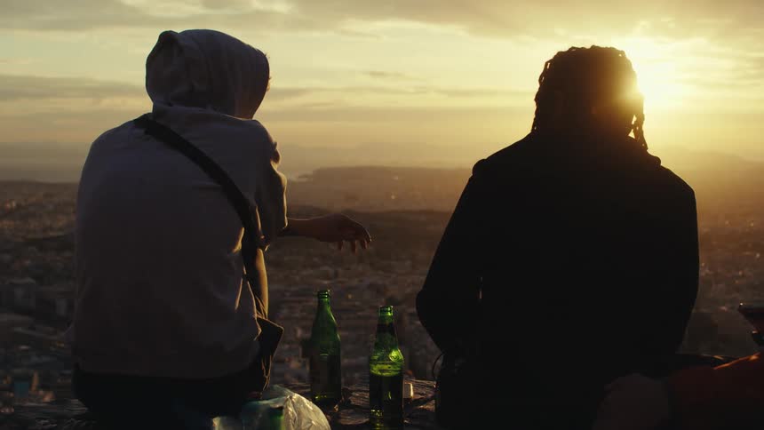 Two friends sitting back to back against a cityscape smoking and holding two bottles of beer