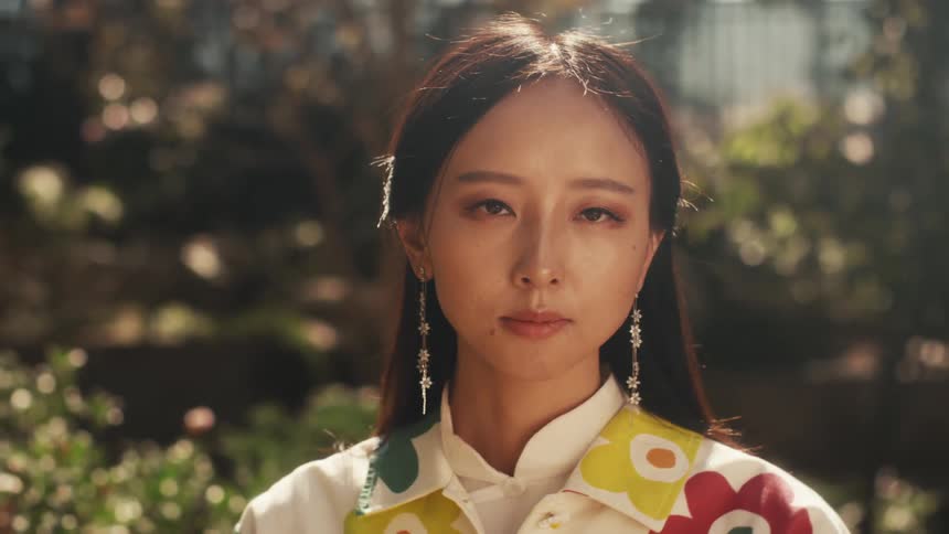 A woman with long hair wearing a white shirt with a colorful floral pattern and long earrings looks at the camera while standing in a park