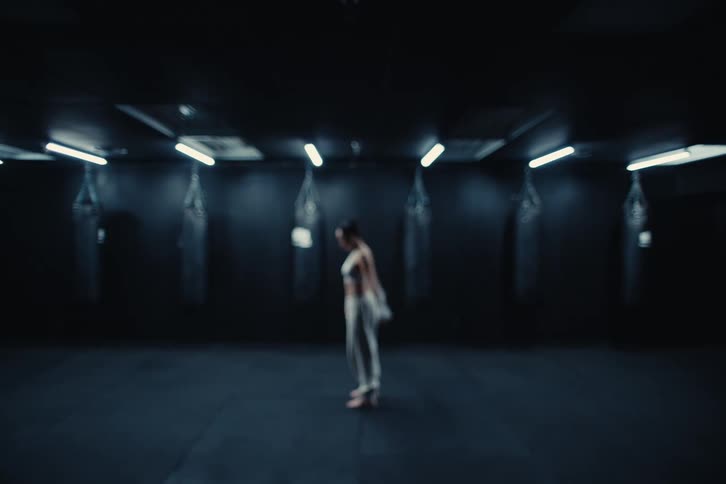 Woman walks through a boxing gym stopping near the body bags before hopping and stretching